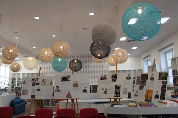 Wnętrze biblioteki na środku stoją cztery sztalugi z planszami wystawy. Nad nimi wiszą kolorowe okrągłe lampy. Z tyłu widać regały z książkami a na górze kolorowe lampy- kliknij aby powiększyć