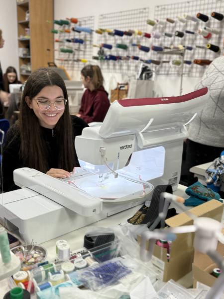 Na zdjęciu uczennica siedząca przy maszynie do haftu. Zdjęcie zostało zrobione podczas warsztatów krawieckich w profesjonalnej pracowni- kliknij aby powiększyć