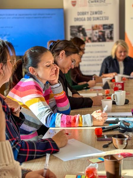 Na zdjęciu uczestniczki warsztatów pn. Radzenie sobie z emocjami. Kobiety siedzą przy stole i piszą na kartkach odpowiedzi na pytanie zadane przez prowadzącą- kliknij aby powiększyć