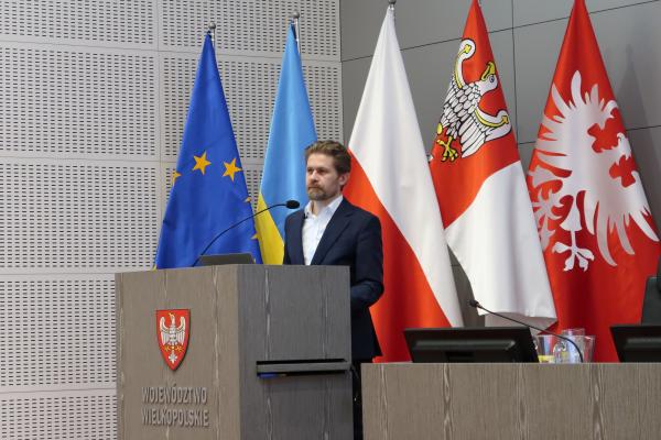 Pan Dyrektor Piotr Łyczko stoi przy mównicy na Sali sesyjnej i wygłasza prezentację dotyczącą nadchodzących zmian w przepisach prawa w związku z implementacją dyrektywy AAQD, a za nim znajduje się pięć flag: Unii Europejskiej, Ukrainy, Polski, Województwa Wielkopolskiego oraz Powstania Wielkopolskiego
- kliknij aby powiększyć