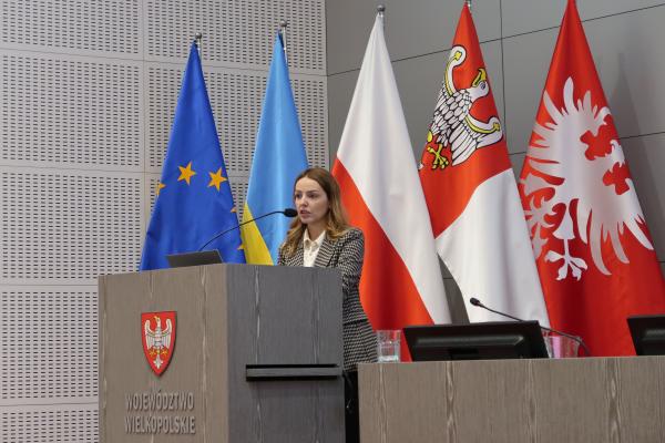 Pani doktor Anna Chlebowska-Styś opowiada przy mównicy o rocznych ocenach jakości powietrza, za nią znajduje się pięć flag: Unii Europejskiej, Ukrainy, Polski, Województwa Wielkopolskiego oraz Powstania Wielkopolskiego
- kliknij aby powiększyć