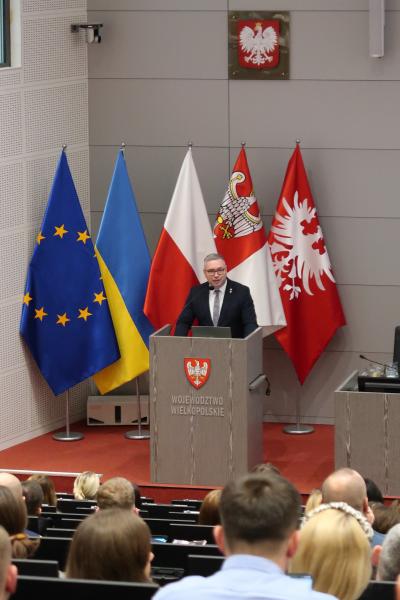 Zdjęcie wykonane z tylnej części sali przedstawiające Członka Zarządu Województwa Wielkopolskiego Jacka Bogusławskiego wygłaszającego przemówienie rozpoczynające wydarzenie- kliknij aby powiększyć