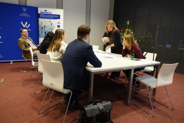 prowadząca pochyla się nad stołem grupy, omawiając notatki z uczestnikami siedzącymi wokół- kliknij aby powiększyć