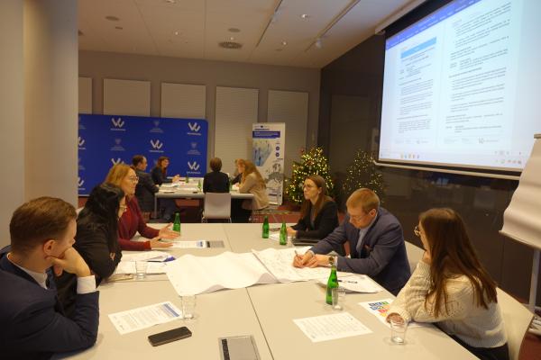 uczestnicy przy głównym stole wypełniają arkusze i rozmawiają, w tle widać kolejną grupę oraz ekran z dokumentem- kliknij aby powiększyć