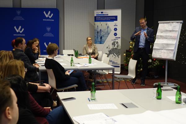 mężczyzna prezentuje wyniki pracy grupy na flipcharcie, pozostali uczestnicy siedzą przy stołach i słuchają- kliknij aby powiększyć