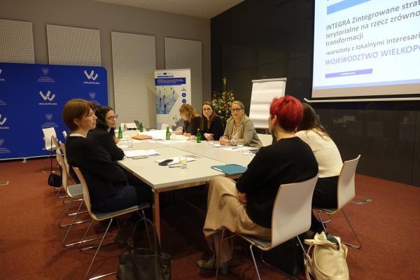 uczestnicy przy głównym stole wypełniają arkusze i rozmawiają, w tle widać kolejną grupę oraz ekran z dokumentem

- kliknij aby powiększyć