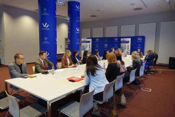 Uczestnicy spotkania siedzą przy długim stole ułożonym w podkowę w nowoczesnej sali konferencyjnej, w której widoczne są niebieskie filary oraz ścianki z logo Wielkopolski i projektu INTEGRA- kliknij aby powiększyć