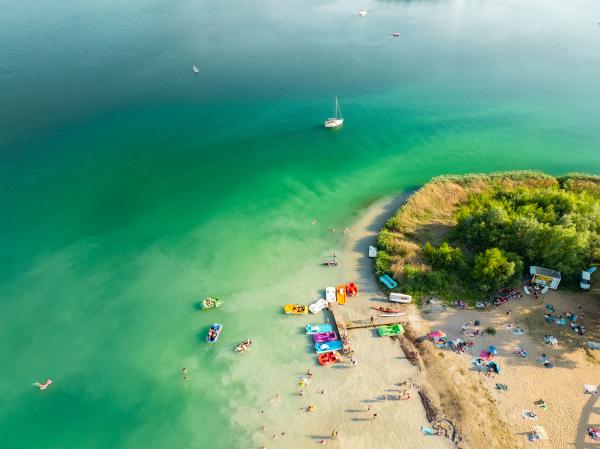 Lotnicze ujęcie turkusowego jeziora z plażą, kąpiącymi się osobami, kolorowymi rowerami wodnymi i żaglówką na spokojnej wodzie- kliknij aby powiększyć